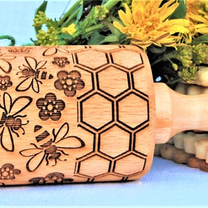 May include: A wooden rolling pin with a honeycomb and bee design. The rolling pin is engraved with the text " Oma Marta".