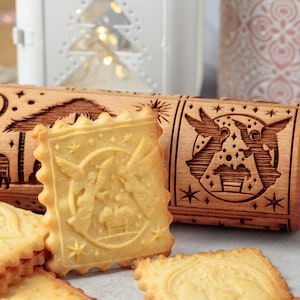 Könnte beinhalten: Ein Holz-Nudelholz mit einem weihnachtlichen Design, das einen Stern, einen Halbmond, einen Baum und einen Engel zeigt. Das Nudelholz wird verwendet, um dekorative Muster auf Kekse zu erstellen. Das Bild zeigt mehrere Kekse, von denen einige mit dem Nudelholz geprägt wurden.