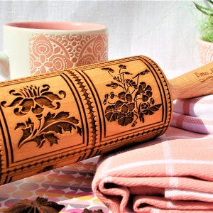 May include: A wooden rolling pin with a floral design engraved on the surface. The rolling pin has a wooden handle with the text "Oma Marta" engraved on it.