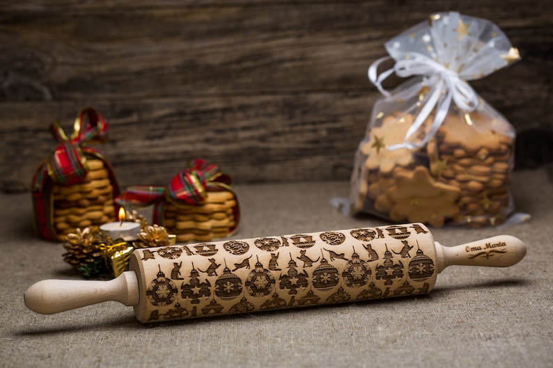 May include: A wooden rolling pin with a Christmas-themed design and the text "Emma Marta" engraved on the handle. The rolling pin is on a burlap surface with Christmas decorations in the background.