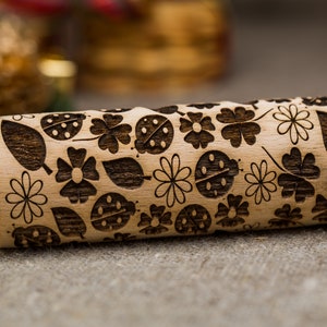 Könnte beinhalten: Ein Holz-Nudelholz mit einem floralen und Marienkäfer-Design. Das Nudelholz ist mit einem Muster aus Blumen, Blättern und Marienkäfern graviert.