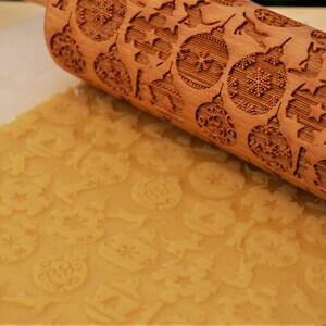 May include: A wooden rolling pin with a Christmas-themed design is rolling over a sheet of cookie dough. The rolling pin has a pattern of snowflakes, ornaments, and stars. The dough is imprinted with the design from the rolling pin.