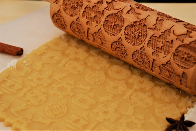 May include: A wooden rolling pin with a Christmas-themed design is rolling over a sheet of cookie dough. The rolling pin has a pattern of snowflakes, ornaments, and stars. The dough is imprinted with the design from the rolling pin.