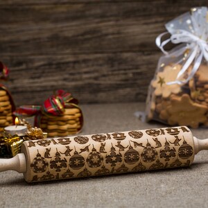May include: A wooden rolling pin with a Christmas-themed design and the text "Emma Marta" engraved on the handle. The rolling pin is on a burlap surface with Christmas decorations in the background.