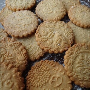May include: A close-up of a variety of homemade cookies with intricate designs. The cookies are light brown and have a slightly rough texture. They are arranged on a brown surface covered in white powder.