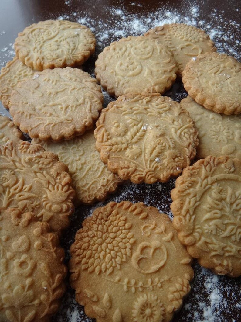 May include: A close-up of a variety of homemade cookies with intricate designs. The cookies are light brown and have a slightly rough texture. They are arranged on a brown surface covered in white powder.