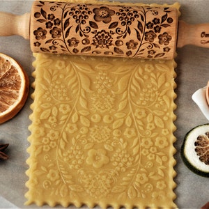 Pode incluir: Um rolo de massa de madeira com um desenho floral gravado na superfície. O rolo é usado para criar um padrão em uma folha de massa. A massa é de cor marrom claro e tem um padrão floral. O rolo tem as palavras "Oma Marta" gravadas na alça.