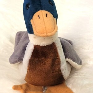 May include: A plush duck toy with a blue head, brown body, and gray wings. The duck has an orange beak and feet, and is set against a white, fluffy background. The toy is soft and cuddly.