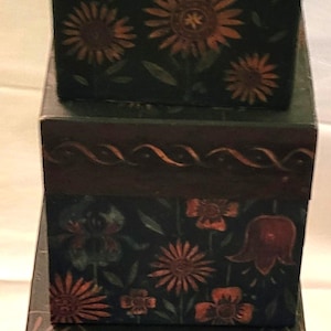 May include: Three stacked, dark green wooden boxes with hand-painted floral designs. The boxes feature sunflowers and other flowers in shades of orange and red. The boxes are arranged in a vertical stack.