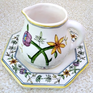 May include: A white ceramic creamer with a yellow, green, and purple floral design. The creamer is sitting on a matching saucer with a yellow, green, and blue floral design.
