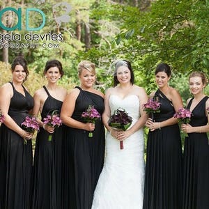 May include: A group of seven women in black dresses stand in a forest setting. The woman in the center is wearing a white wedding dress and a veil. The other women are bridesmaids and are holding bouquets of purple flowers. The image includes the text "Angela DeVries Photography.com".
