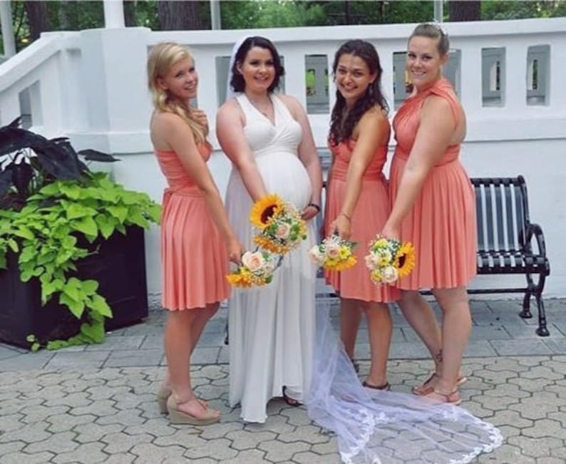 Puede incluir: Una novia con un vestido blanco posa con tres damas de honor con vestidos color coral. Cada mujer sostiene un ramo de flores, incluyendo girasoles. La escena sugiere una boda al aire libre.
