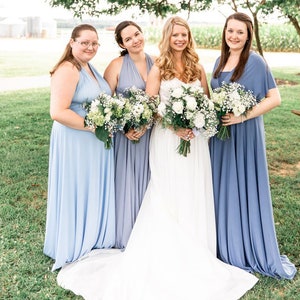 May include: Four women in a field, three wearing light blue maxi dresses and one wearing a white wedding dress. They are holding bouquets of white flowers.