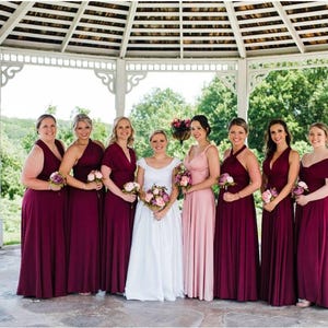 May include: A group of women, including a bride, pose for a photo in a gazebo. The bridesmaids wear long, burgundy dresses and hold bouquets. The bride wears a white wedding dress and holds a bouquet. The setting is outdoors with trees in the background.