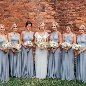 May include: Seven women in elegant, floor-length, light gray dresses pose in front of a brick wall. Each woman holds a bouquet of flowers. The bride wears a white dress with a floral bouquet.