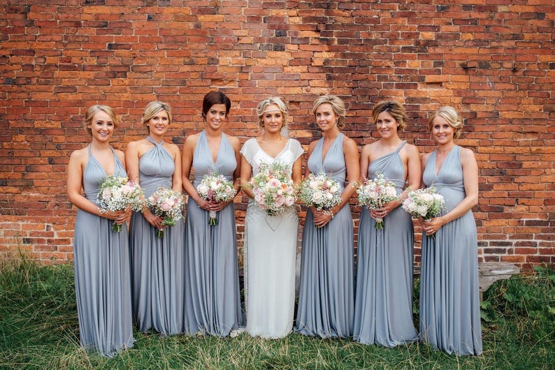 May include: Seven women in elegant, floor-length, light gray dresses pose in front of a brick wall. Each woman holds a bouquet of flowers. The bride wears a white dress with a floral bouquet.