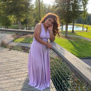 May include: A light purple halter-style maxi dress. The dress features a flowing skirt and is made of a soft, draping fabric. The woman is leaning on a wooden railing on a bridge, with trees and a lake in the background.