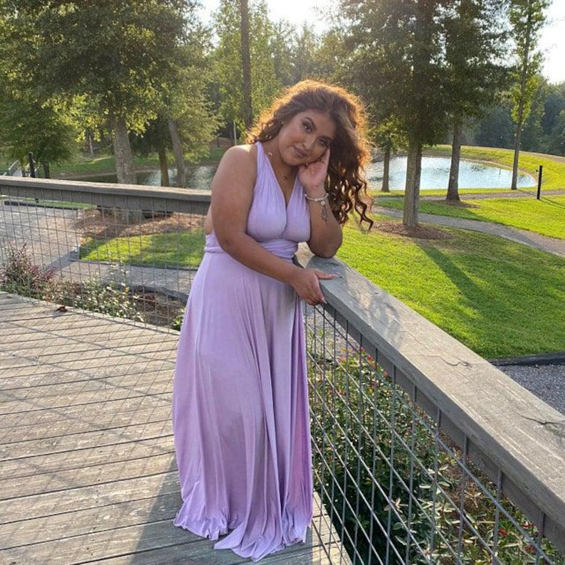 May include: A light purple halter-style maxi dress. The dress features a flowing skirt and is made of a soft, draping fabric. The woman is leaning on a wooden railing on a bridge, with trees and a lake in the background.