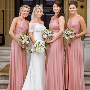 May include: A bride in a white wedding dress with lace sleeves and three bridesmaids in dusty rose-colored dresses. Each woman holds a bouquet of flowers. The wedding party is standing in front of a dark wooden door.