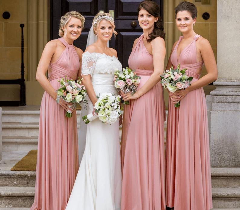 May include: A bride in a white wedding dress with lace sleeves and three bridesmaids in dusty rose-colored dresses. Each woman holds a bouquet of flowers. The wedding party is standing in front of a dark wooden door.