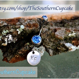 May include: A silver belly button ring with two blue crystal stones and a charm with the initials "KE" engraved on it.