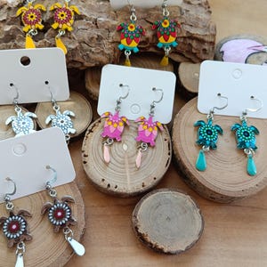 May include: A selection of turtle-shaped earrings in various colours, displayed on wooden slices. The earrings feature floral patterns and dangling accents. The earrings are presented on white display cards.