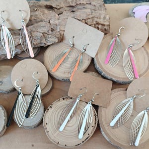 Può includere: Una collezione di orecchini fatti a mano esposti su fette di legno e cartoncini. Gli orecchini presentano un design traforato con una varietà di colori, tra cui rosa, arancione, nero e bianco. Gli orecchini sono sospesi da ganci color argento.