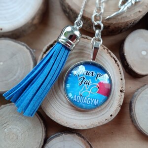 Puede incluir: Collar con colgante plateado con un gráfico azul y blanco. El colgante tiene el texto "peux pas Jai AQUAGYM" en blanco. Una borla azul cuelga de la cadena. El collar se exhibe sobre una superficie de madera.