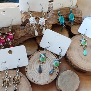 May include: A selection of turtle-shaped earrings in various colours and patterns, including red, white, blue, green, and leopard print. The earrings are displayed on wooden slices and a textured wooden surface. Each pair is attached to a white earring display card.