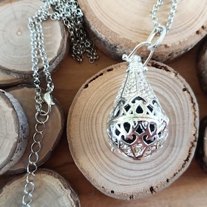 Può includere: Una collana con pendente color argento con un design a gabbia a forma di goccia, incrostata di piccoli cristalli trasparenti. Il pendente pende da una delicata catena. La collana è appoggiata su una superficie di legno.