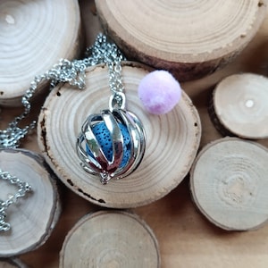 May include: Silver-colored essential oil diffuser necklace with a blue stone inside a cage-like pendant. The necklace is on a wooden surface with wood slices and a small, light purple pom-pom.