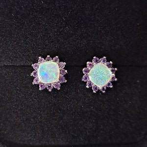 May include: A pair of stud earrings featuring square, iridescent opal center stones surrounded by small, round, purple amethyst stones. The earrings are set in a silver metal and rest on a black surface.