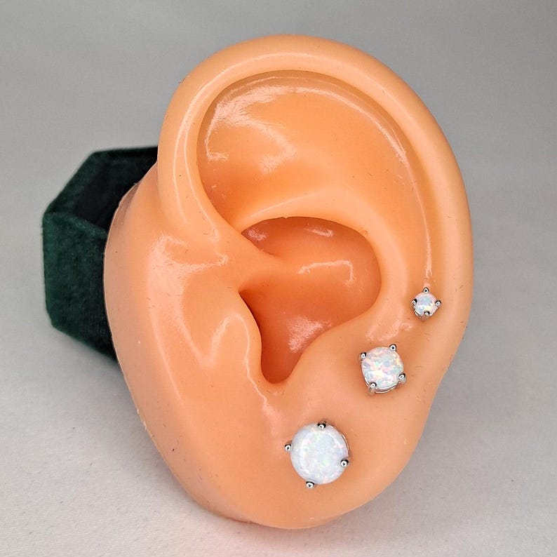 Puede incluir: Tres pendientes de bot&oacute;n de &oacute;palo blanco de diferentes tama&ntilde;os se muestran en un modelo de oreja color melocot&oacute;n. Los pendientes est&aacute;n engarzados en metal plateado. El modelo de oreja est&aacute; colocado frente a una caja de terciopelo verde oscuro.