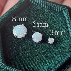 Puede incluir: Tres pendientes de bot&oacute;n de &oacute;palo en tama&ntilde;os de 8 mm, 6 mm y 3 mm, exhibidos sobre una superficie de terciopelo verde oscuro. Los pendientes tienen un engaste plateado y una piedra de &oacute;palo redonda e iridiscente. El texto "8mm", "6mm" y "3mm" es visible.