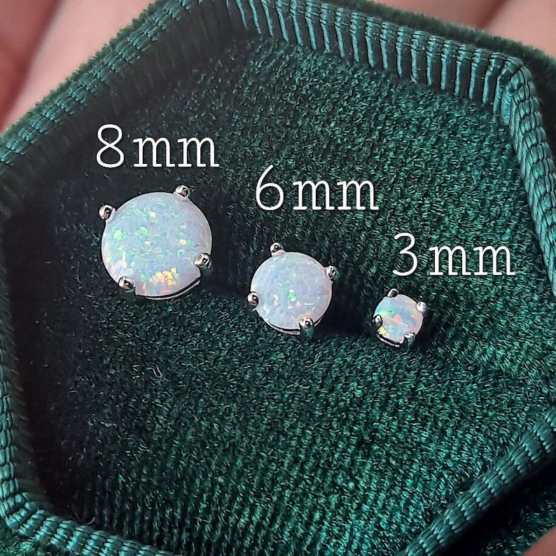 Puede incluir: Tres pendientes de bot&oacute;n de &oacute;palo en tama&ntilde;os de 8 mm, 6 mm y 3 mm, exhibidos sobre una superficie de terciopelo verde oscuro. Los pendientes tienen un engaste plateado y una piedra de &oacute;palo redonda e iridiscente. El texto "8mm", "6mm" y "3mm" es visible.