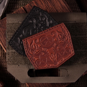 Puede incluir: Dos parches rectangulares de cuero con diseños de cráneos de ciervo en relieve y patrones de remolinos intrincados. Un parche es negro y el otro es marrón rojizo. Ambos están colocados sobre una base de tela verde oliva con correas horizontales.