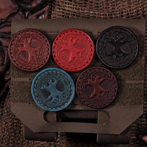 May include: Five circular leather patches featuring an embossed tree design with intricate knotwork borders. The patches are displayed in various colors including red, black, blue, and brown, and are attached to a rectangular olive green fabric panel.