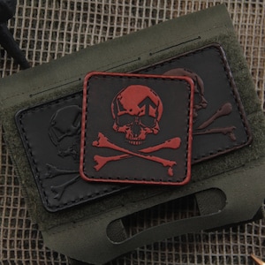 May include: Close-up of a tactical pouch with two skull and crossbones patches. The patches are black and red, with a skull and crossbones design. The pouch is olive green.