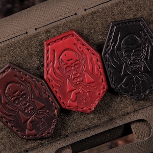 May include: Three coffin-shaped patches in dark brown, bright red, and black leather. Each patch features an embossed graphic of a vampire with pointed ears and a high-collared cape, with visible stitching along the edges of the leather shapes.