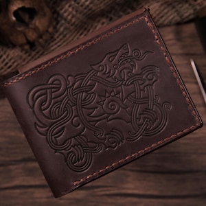 Può includere: Un portafoglio bifold in pelle marrone scuro con un intricato design a intreccio in rilievo raffigurante una creatura simile a un lupo sulla copertina anteriore. Il portafoglio è realizzato con cuciture marrone scuro visibili lungo i bordi e ha una finitura opaca.