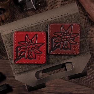 May include: Two square leather patches featuring an embossed edelweiss flower design. One patch is bright red and the other is a darker reddish-brown. Both patches are attached to an olive green fabric panel with horizontal webbing slots.
