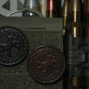 May include: Two leather patches with a tree design. The patches are brown and black and have a stitched border. The patches are attached to a green fabric background.