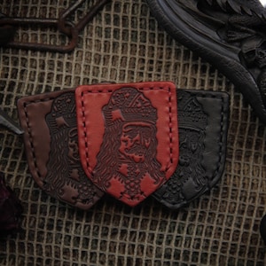 May include: Three leather patches in shades of brown, red, and black, each with an embossed image of a crowned figure. The patches are shield-shaped with stitched edges. The background is a textured, woven material.