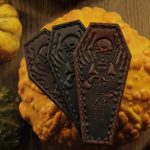 May include: Three leather coffin-shaped accessories in shades of brown and black, featuring embossed vampire designs. The accessories are displayed on a textured orange gourd, with additional gourds in the background. The overall theme is Halloween or gothic.