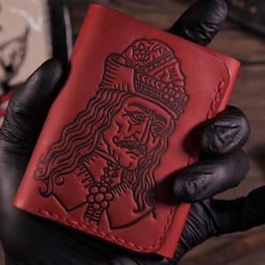 Könnte beinhalten: Rotes Lederportemonnaie mit einem detaillierten geprägten Porträt einer historischen Figur. Das Portemonnaie hat ein detailliertes Design und wird von einer Person mit schwarzen Handschuhen gehalten. Das Portemonnaie hat eine satte rote Farbe.