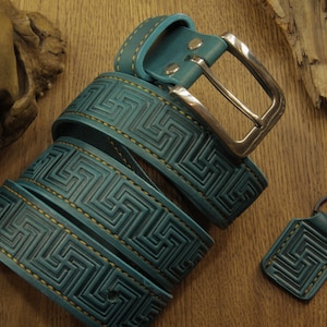May include: Teal leather belt with a geometric pattern and a silver buckle. The belt is accompanied by a matching keychain. The leather is stitched with a contrasting gold thread. The belt is rolled up on a wooden surface.