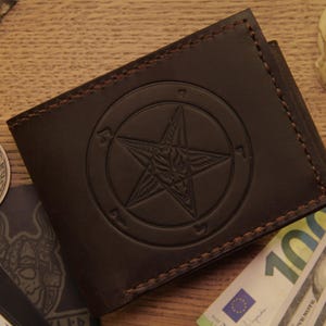 Könnte beinhalten: Dunkelbraune Ledergeldbörse mit einem geprägten Pentagramm-Design auf der Vorderseite. Die Geldbörse hat braune Nähte und liegt neben einem Totenkopf, Münzen und Bargeld. Es ist ein zweifach gefalteter Stil.