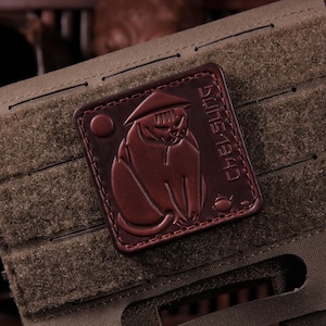 May include: A brown leather patch with a cat in a hat and the text "CHELSUNG 1943". The square patch is stitched onto a green fabric with a hook and loop fastener.