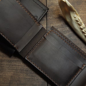 May include: Open, dark brown leather wallet with visible stitching and card slots. The wallet is displayed open, revealing its interior. A second wallet is partially visible in the background. A bone fragment is in the background.