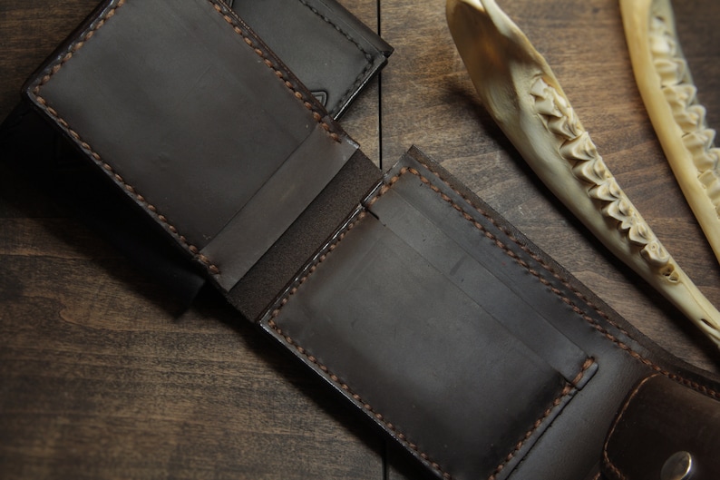 May include: Open, dark brown leather wallet with visible stitching and card slots. The wallet is displayed open, revealing its interior. A second wallet is partially visible in the background. A bone fragment is in the background.
